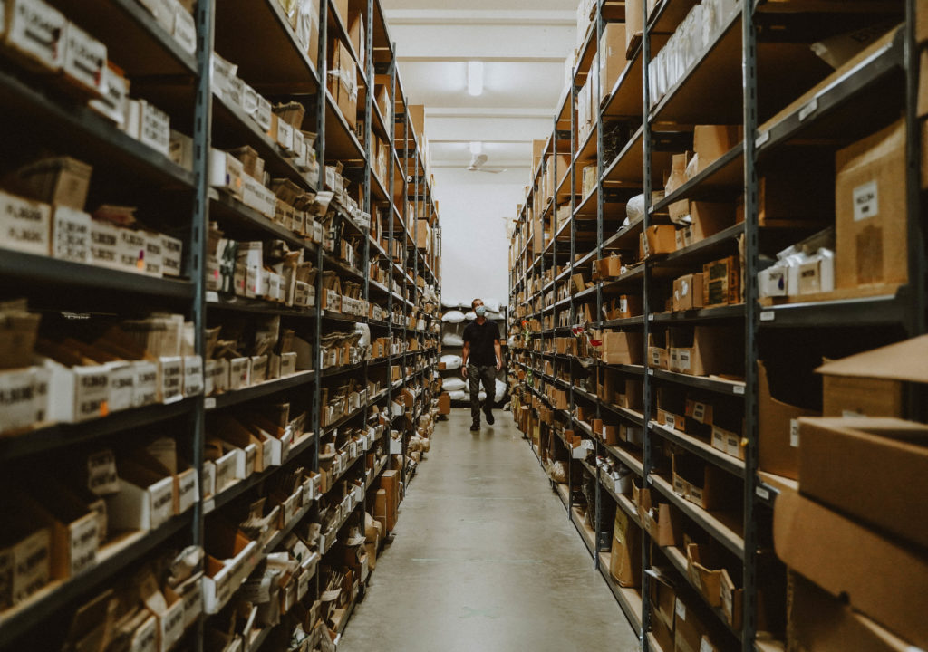 A man is seen in the middle of two large shelves full of seeds and other products ready to be shipped.
